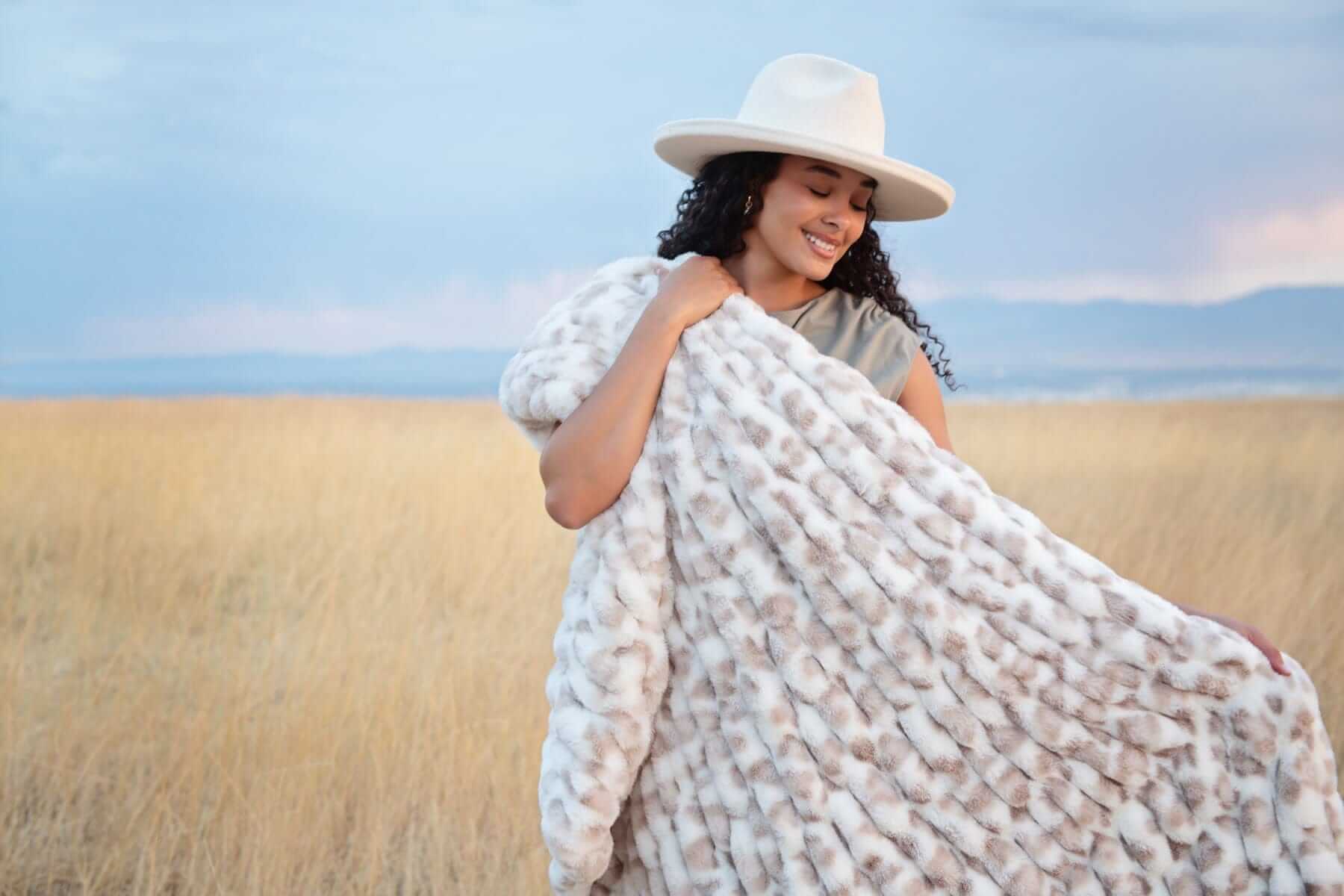Woman with a white hat and a snow leopard-print faux fur blanket in a field.