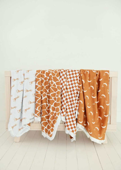 Four Minky Couture baby blankets in warm tan and white patterns draped over a wooden crib railing.