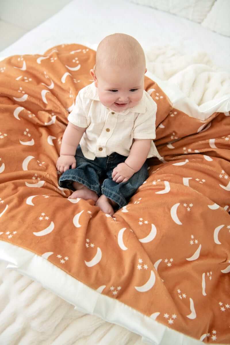 A baby sitting on the Shining Moon Copper Blanket with cream half-moons against a warm rusty brown background.