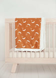 Shining Moon Copper Blanket draped over a crib, featuring cream moons and stars on a warm rusty brown background.