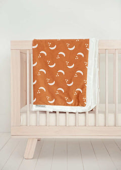 Shining Moon Copper Blanket draped over a crib, featuring cream moons and stars on a warm rusty brown background.