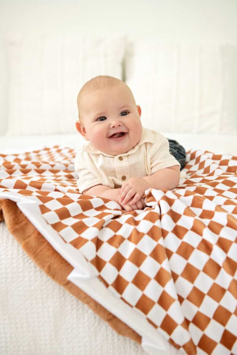 A happy baby lying on a Minky Couture Checkered Copper Blanket with a hazelnut brown and cream pattern.