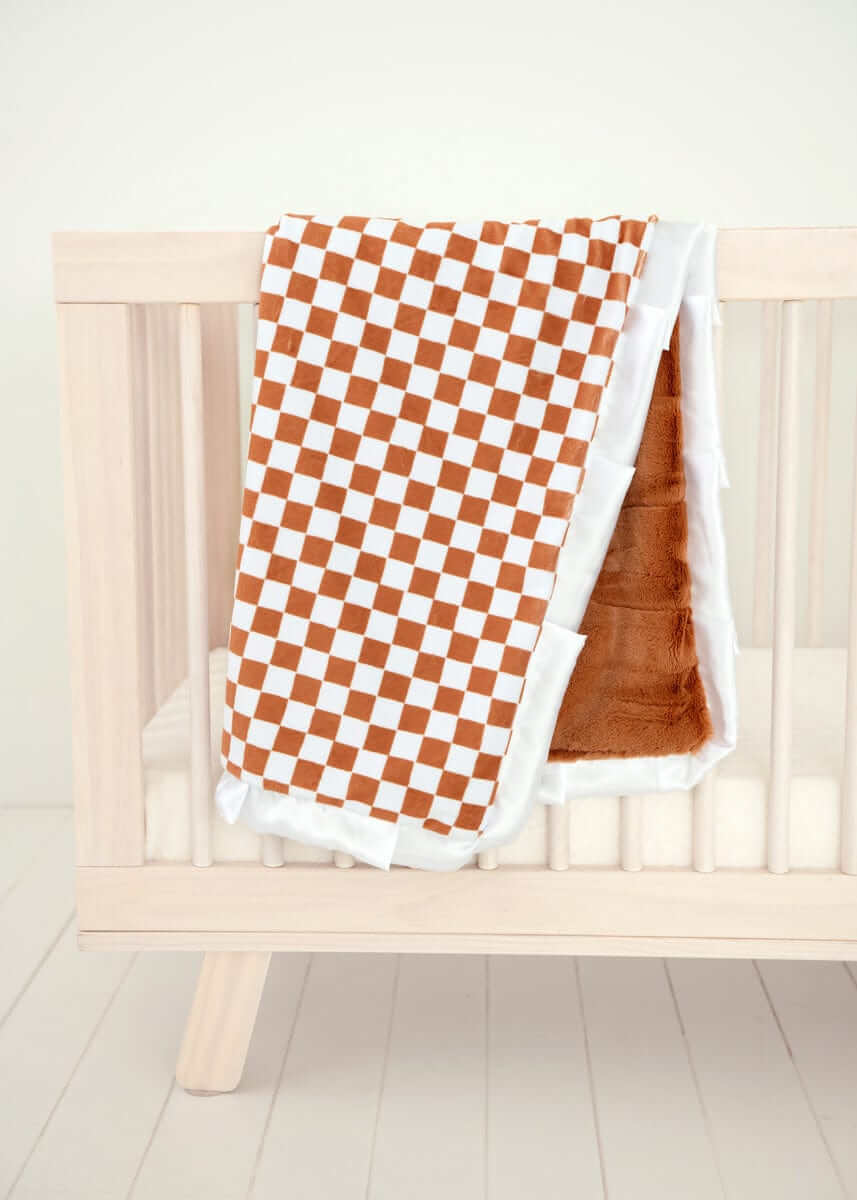 Checkered Copper Blanket draped over a wooden crib, featuring a hazelnut brown and cream checkered pattern with plush backing.
