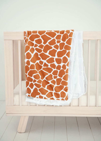 Minky Couture Giraffe Blanket displayed on crib with classic giraffe pattern in warm rusty brown and cream colors.