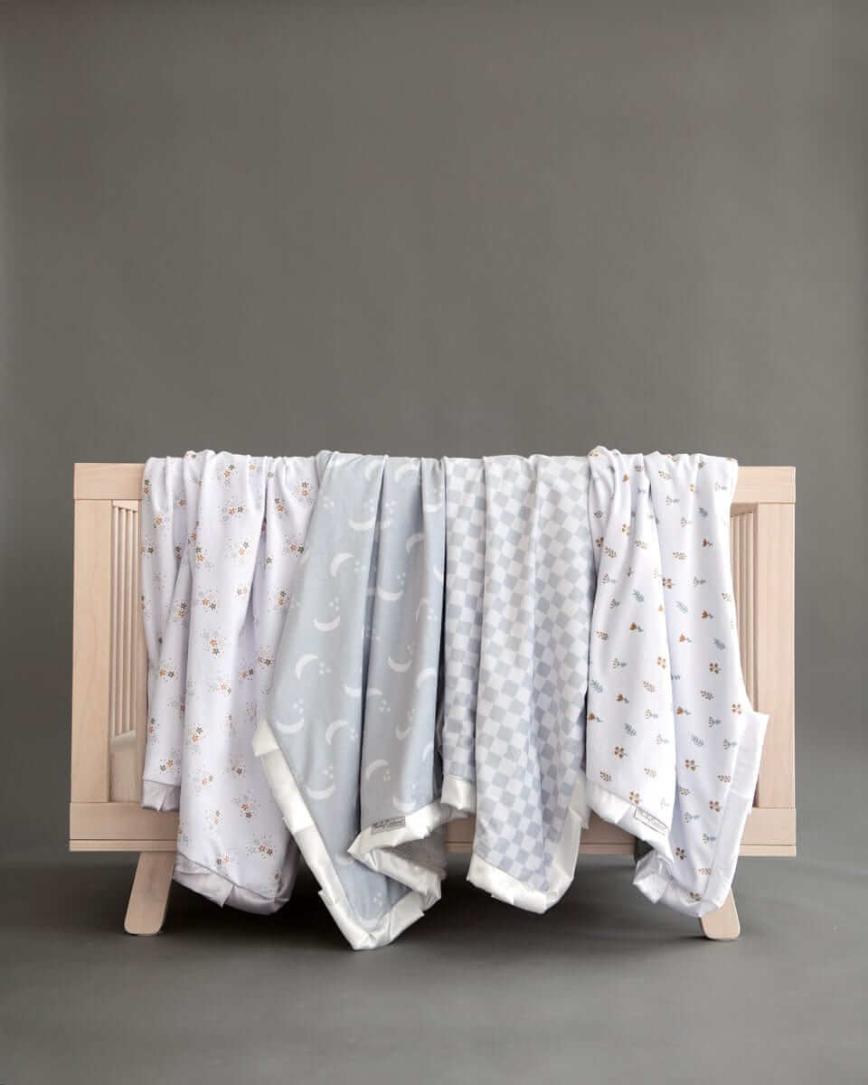 Four neutral baby Minky Couture Blankets draped over wooden crib in soft gray and white patterns.