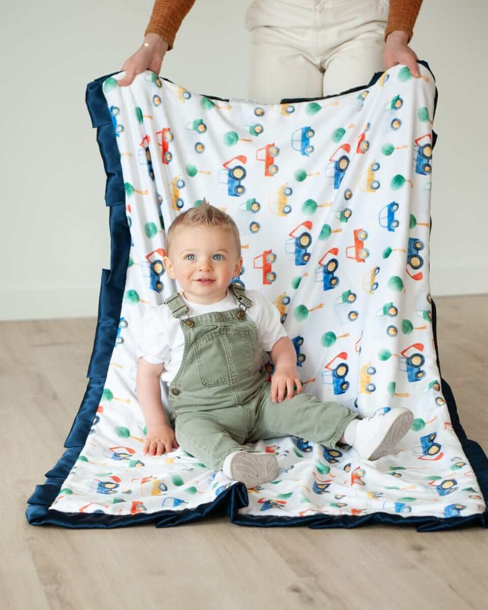 A happy baby sitting on a Cars Blanket featuring colorful cars and tractors, surrounded by a plush navy satin ruffle.
