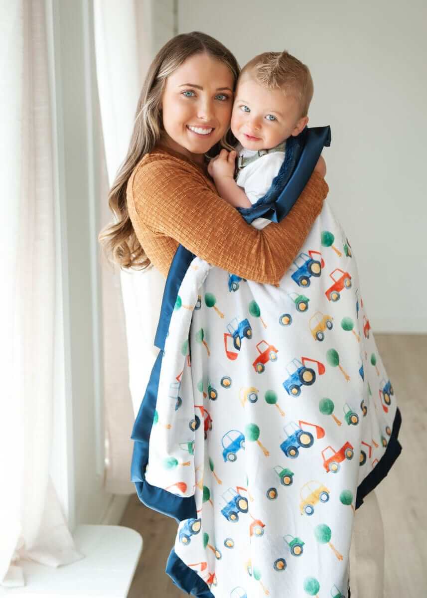 Mom and child snuggled in a colorful Cars Blanket featuring cars and tractors with a navy plush backing.
