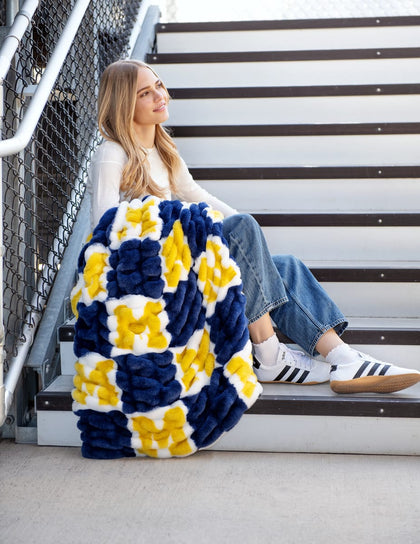 University of Michigan Wolverines Blanket draped on a young woman sitting on stairs, showcasing the vibrant blue and yellow pattern.