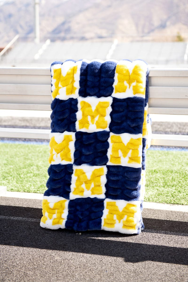 University of Michigan Wolverines Blanket displayed on a bench with a vibrant design of M's in blue and yellow.