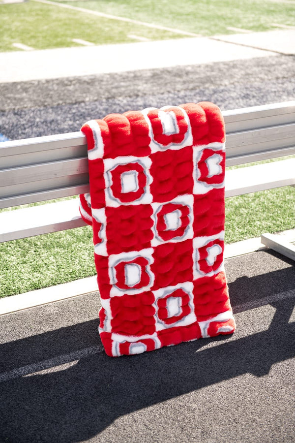 Ohio State Buckeyes Hugs Blanket displayed on a bench at a football field, showcasing its vibrant red colors and cozy design.