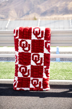 University of Oklahoma Sooners Blanket displayed on a bench at a sports field, showcasing its bold red and white design.
