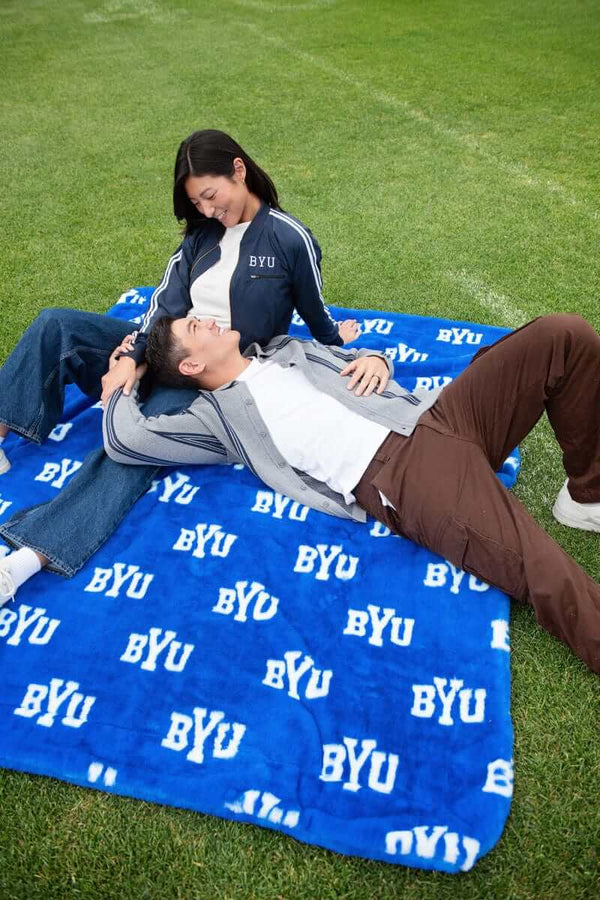 BYU Cougars Blanket featuring a couple relaxing on a grass field with the iconic BYU logo on a blue background.