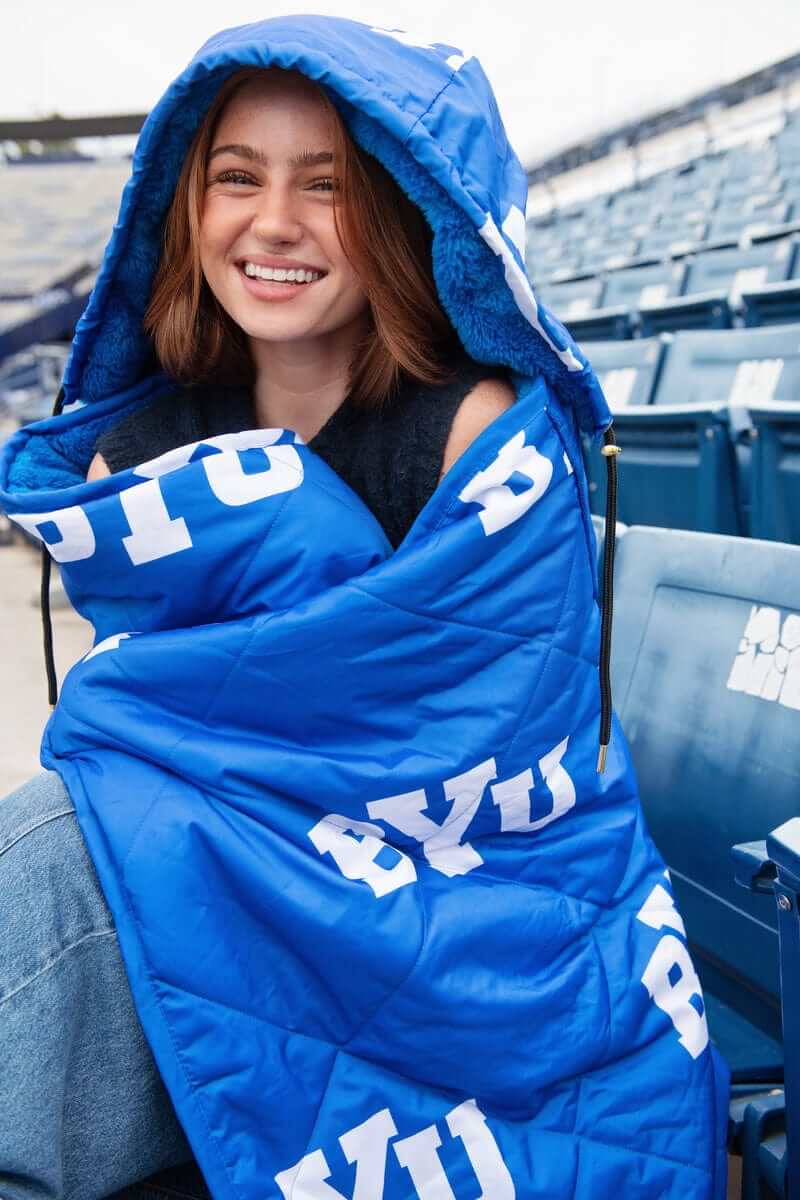 Woman wearing the Minky Couture BYU Cougars Hooded Stadium Blanket in a stadium setting, showcasing team spirit.