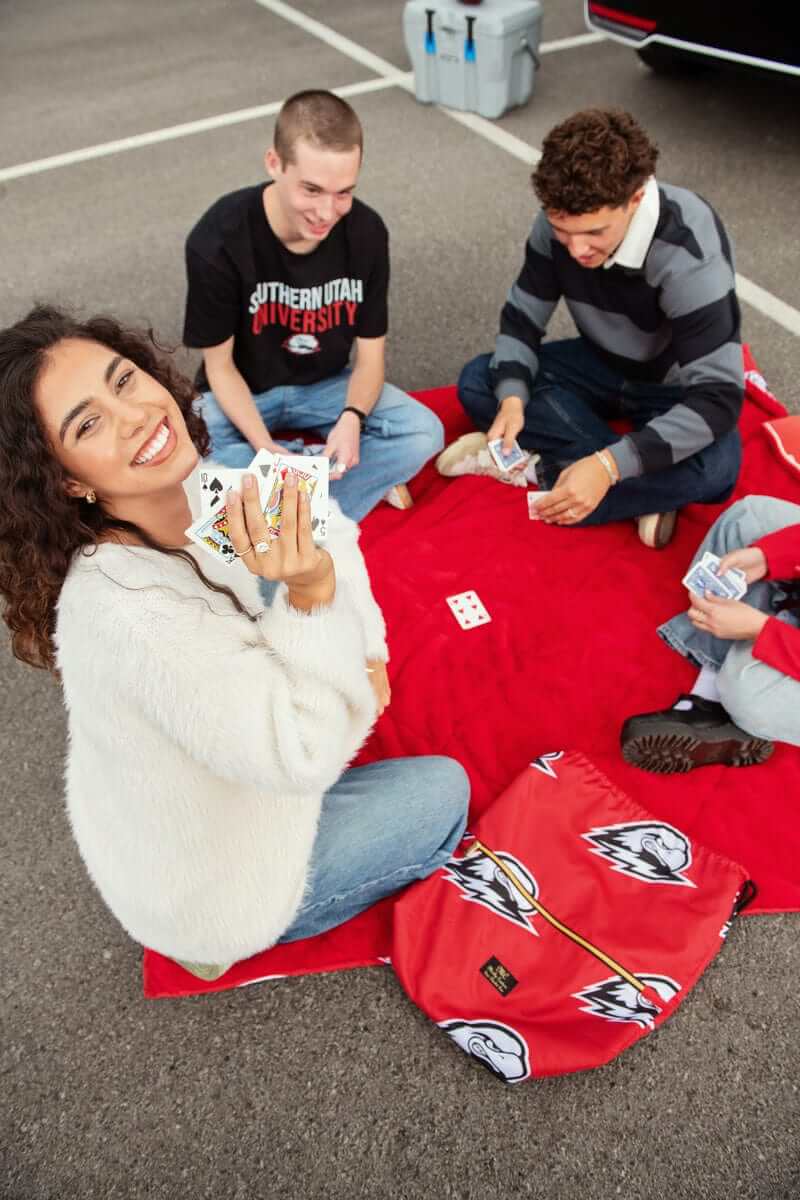 Friends playing cards on a red Southern Utah University Minky Couture blanket in a parking lot.