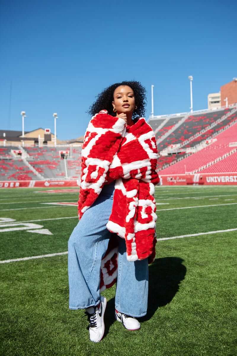 Woman wearing red and white University of Utah blanket coat on football field with stadium seats behind.