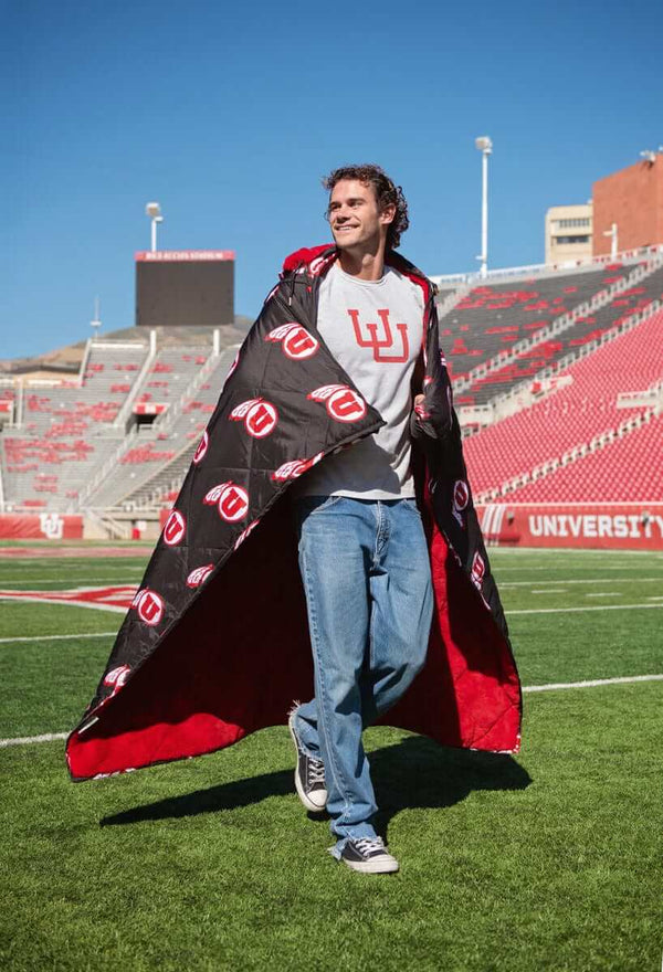 Man walking on football field wearing black and red University of Utah Hooded blanket like a cape in stadium.