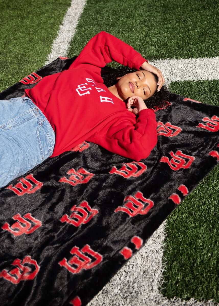 Woman lying on football field wrapped in University of Utah black plush blanket with red U logo pattern.
