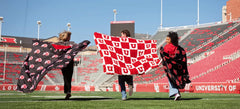 Three fans running on football field holding Minky Couture red and black University of Utah logo blankets in stadium.