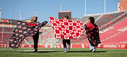Three fans running on football field holding Minky Couture red and black University of Utah logo blankets in stadium.