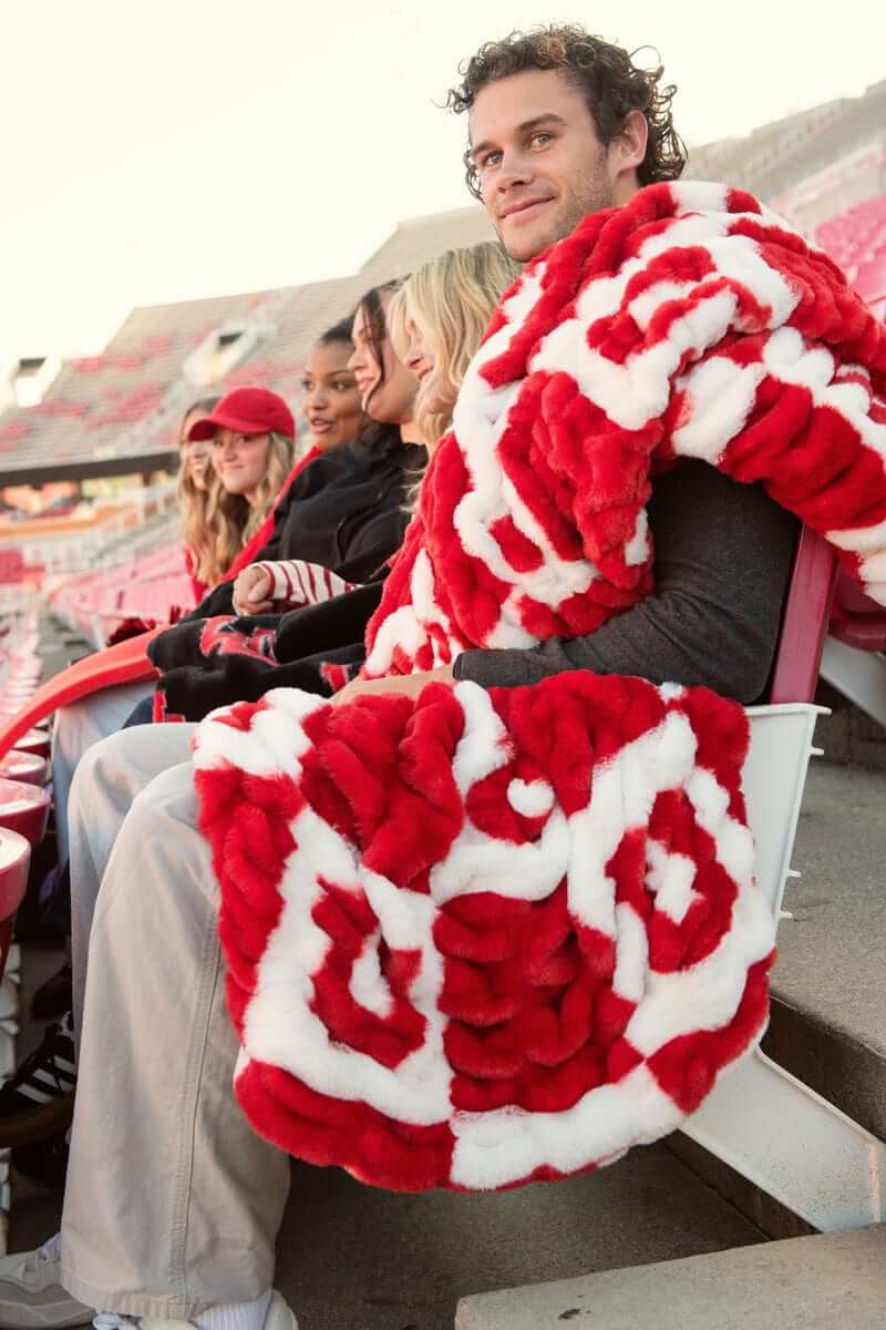 Man wrapped in Minky Couture red and white University of Utah blanket sitting in stadium seats with friends.
