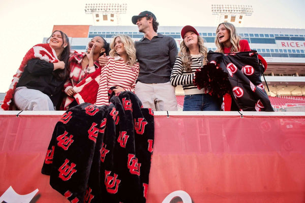 Group of fans at stadium railing holding black and red U logo blankets, cheering together.
