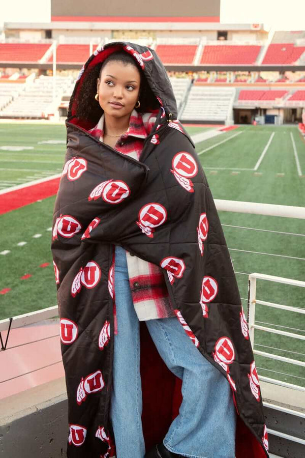 Woman wrapped in black University of Utah Utes Hooded Stadium Blanket at football stadium stands wearing plaid shirt and jeans.