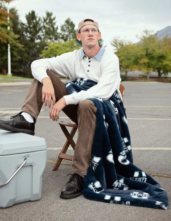 Navy Utah State Aggies blanket draped over seated man at tailgate in parking lot.