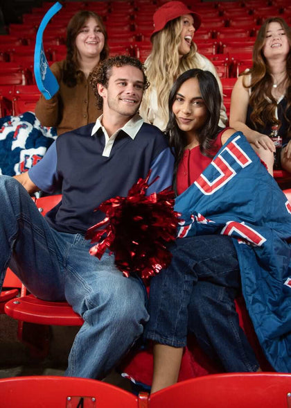 Couple sitting in red stadium seats wrapped in blue UT blanket with friends cheering behind.