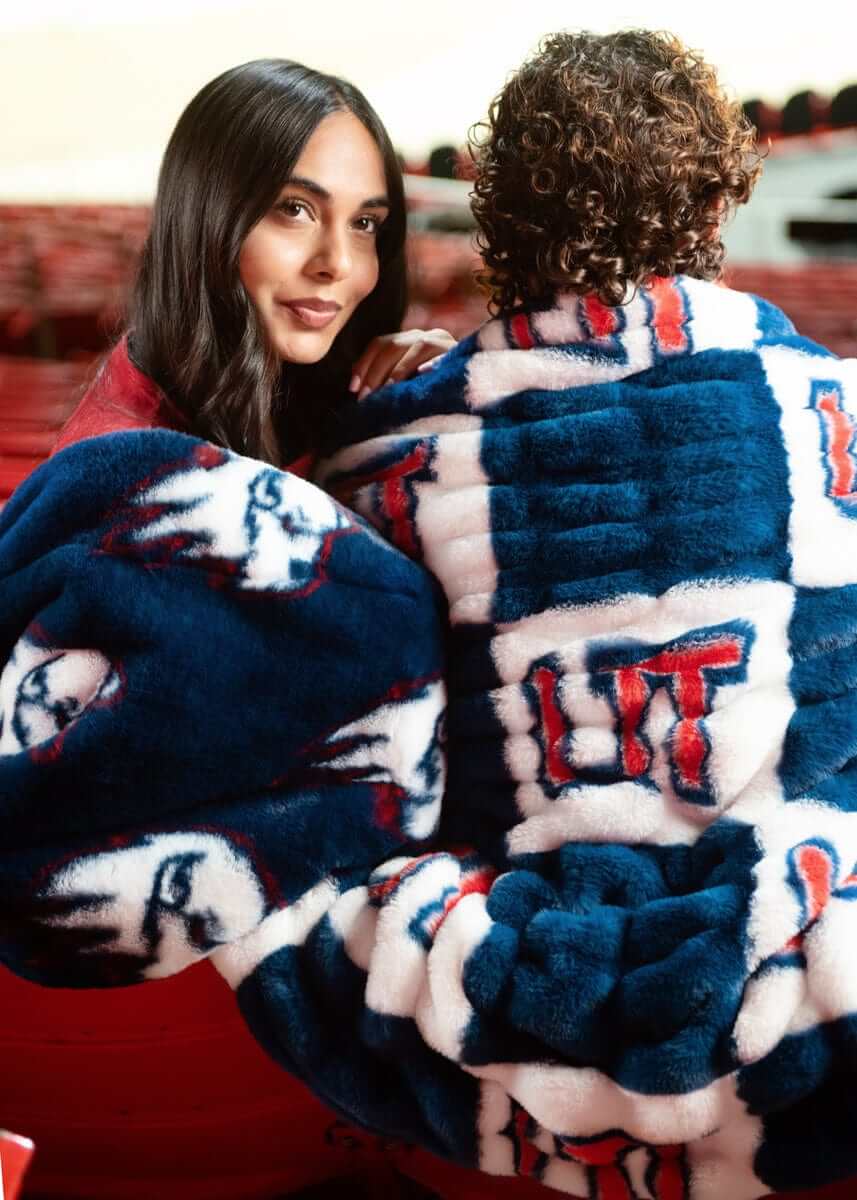 Two people wrapped in blue and white UT Tech Trailblazers  plush blanket with eagle logo indoors.