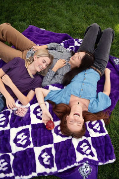 Three friends relaxing on a purple Wildcats blanket on grass, laughing together outdoors.