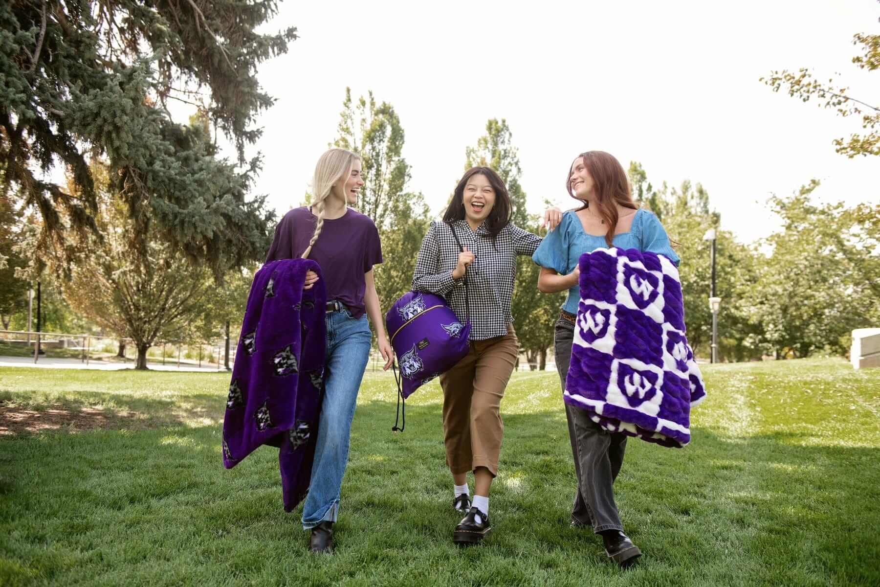 Three women walking on grass with purple Weber State Wildcats blankets and matching drawstring bag.
