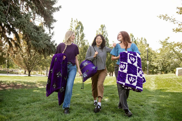 Three women walking on grass with purple Weber State Wildcats blankets and matching drawstring bag.