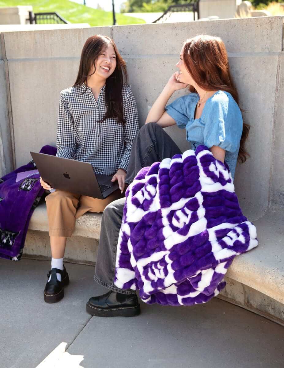 Two women sitting on campus steps with a purple Wildcats checkered blanket and laptop.