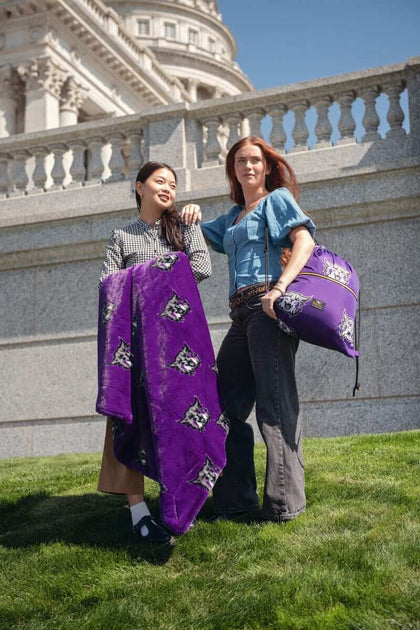 Two women holding purple Weber State Wildcats blanket and pillow on grass in front of historic building.