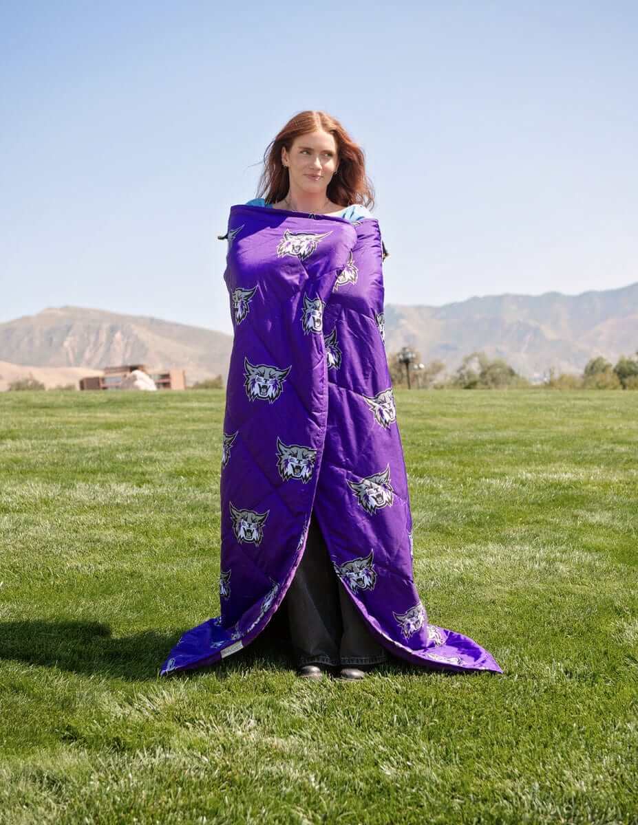Woman wrapped in purple Weber State Wildcats blanket standing on grassy field with mountains in background.