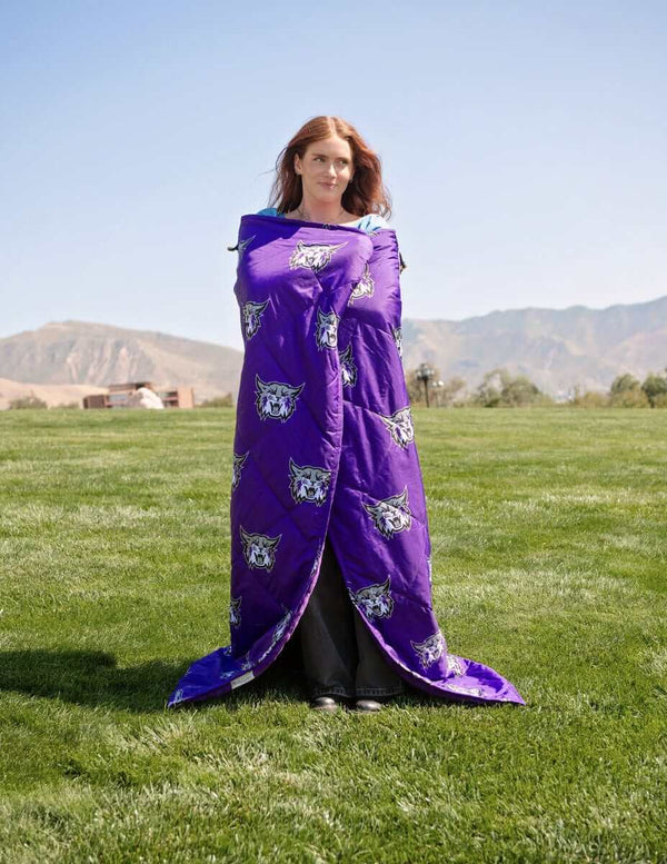 Woman wrapped in purple Weber State Wildcats blanket standing on grassy field with mountains in background.