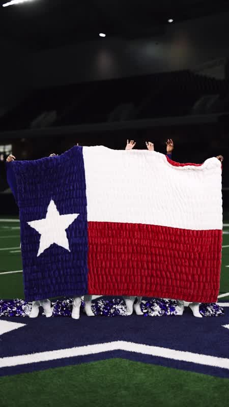 A large Texas flag blanket held up by three people standing in white cowboy boots, with blue and white pom-poms on a football field.