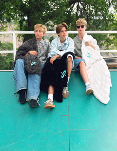 Three young men sitting together on a teal outdoor surface, each wearing Minky Couture Dude Perfect Blanket throw in black, gray, and cream with turquoise logo accents. Trees and a white railing are visible in the background.