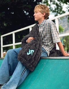 A young man sits on a green ramp wearing a plaid shirt and jeans, holding a Minky Couture Dude Perfect Hugs Charcoal blanket with a light blue Dude Perfect logo. Trees and a white fence are visible in the background.