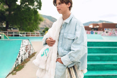 A young man stands at an outdoor skatepark wearing a light blue vertically striped button-up shirt, a Minky Couture Dude Perfect Hugs Cream blanket draped over his arm.