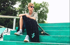 A person sitting on bright green outdoor stairs, wearing a light gray shirt, dark shorts, and white sneakers, with a black hugs black Minky Couture Dude Perfect blanket over their lap and a skateboard beside them.