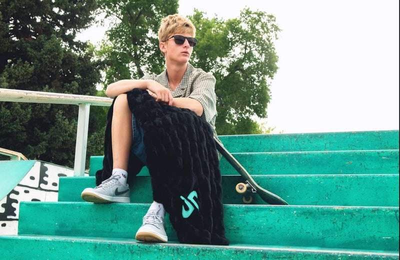A person sitting on bright green outdoor stairs, wearing a light gray shirt, dark shorts, and white sneakers, with a black hugs black Minky Couture Dude Perfect blanket over their lap and a skateboard beside them.
