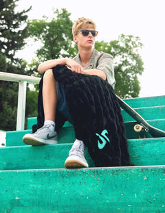 A young man wearing sunglasses sits on green outdoor bleachers, holding a black hugs blanket with a turquoise Dude Perfect logo. He is wearing a checkered shirt, denim shorts, and gray sneakers.