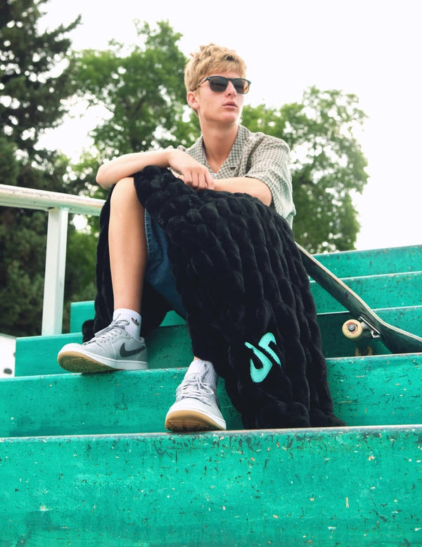 A young man wearing sunglasses sits on green outdoor bleachers, holding a black hugs blanket with a turquoise Dude Perfect logo. He is wearing a checkered shirt, denim shorts, and gray sneakers.