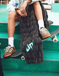 A person sitting on green concrete bleachers with a skateboard, wrapped in a chunky  Minky Couture Dude Perfect Hugs Charcoal blanket featuring a light blue  Dude Perfect logo.