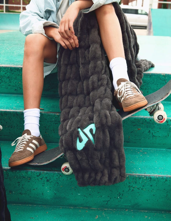 A person sitting on green concrete bleachers with a skateboard, wrapped in a chunky  Minky Couture Dude Perfect Hugs Charcoal blanket featuring a light blue  Dude Perfect logo.
