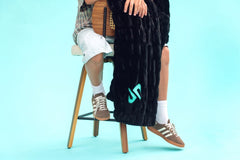 A person seated on a wooden stool against a light blue studio background, draped in a black Minky Couture Dude Perfect Hugs  blanket with a turquoise Dude Perfect logo. The person wears white shorts, brown sneakers, white socks. 