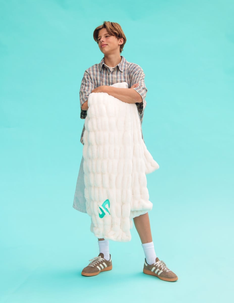 A model stands against a solid turquoise background holding a fluffy white Minky Couture Dude Perfect Hugs Cream blanket with a green  Dude Perfect logo. The model is wearing a plaid shirt, white socks, and brown sneakers.
