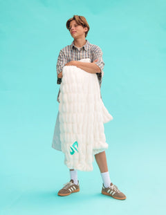 A model stands against a solid turquoise background holding a fluffy white Minky Couture Dude Perfect Hugs Cream blanket with a green  Dude Perfect logo. The model is wearing a plaid shirt, white socks, and brown sneakers.