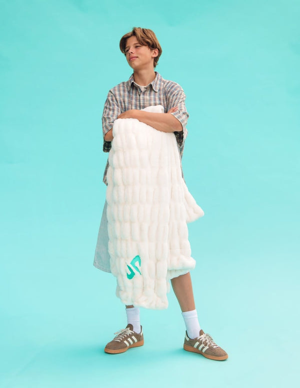A model stands against a solid turquoise background holding a fluffy white Minky Couture Dude Perfect Hugs Cream blanket with a green  Dude Perfect logo. The model is wearing a plaid shirt, white socks, and brown sneakers.